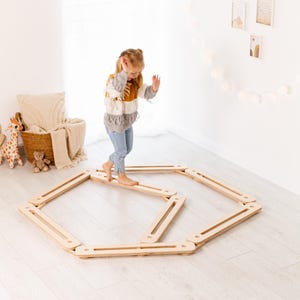 May include: A child balances on a wooden balance board in a bright room. The balance board is made of light-coloured wood and is arranged in a hexagonal shape. The child is wearing a colourful sweater and jeans.