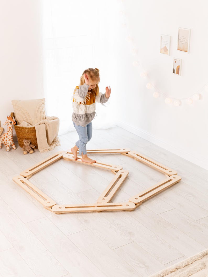 May include: A child balances on a wooden balance board in a bright room. The balance board is made of light-coloured wood and is arranged in a hexagonal shape. The child is wearing a colourful sweater and jeans.