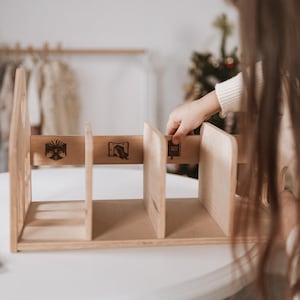 May include: A light-coloured wooden bookshelf with three compartments. The bookshelf has decorative etched designs on the front. A person's hand is reaching towards the bookshelf. The bookshelf is on a white table.
