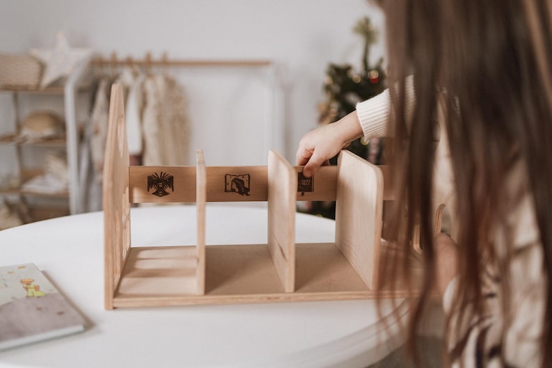 May include: A light-coloured wooden bookshelf with three compartments. The bookshelf has decorative etched designs on the front. A person's hand is reaching towards the bookshelf. The bookshelf is on a white table.