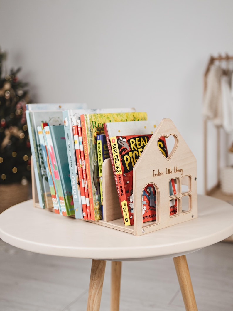 May include: A wooden bookshelf shaped like a house, filled with colorful children's books. The bookshelf has a heart-shaped cutout and the text "Emilia's Little Library". The books are on a light-colored round table.