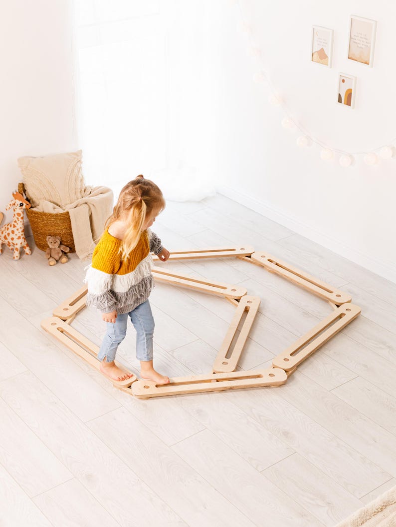 May include: A child in a yellow, white, and grey sweater and blue jeans walks on a wooden balance beam. The balance beam is made of light-coloured wood and is arranged in an octagonal shape on a light-coloured wooden floor. Toys and decor are in the background.