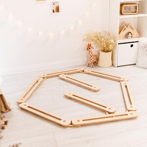 May include: A wooden balance board set for children, comprising a hexagonal frame and several straight planks. The natural wood colour complements the neutral-toned room, which includes a giraffe plush toy and decorative wall art. The set is designed for indoor play.