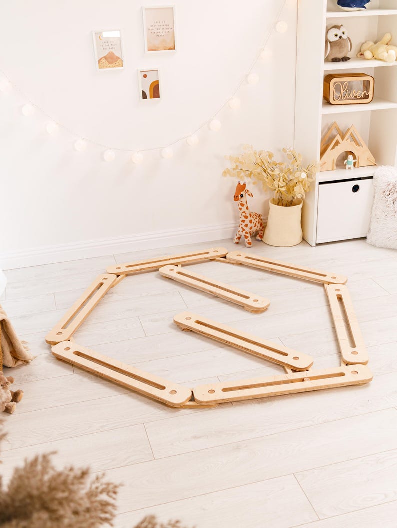 May include: A wooden balance board set for children, comprising a hexagonal frame and several straight planks. The natural wood colour complements the neutral-toned room, which includes a giraffe plush toy and decorative wall art. The set is designed for indoor play.