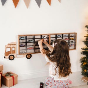 Op de afbeelding: Een houten vrachtwagenvormige displaykast met meerdere compartimenten, met speelgoedauto's. De kast is aan een witte muur bevestigd, met een kind dat met het speelgoed speelt. De tekst "Tom's Garage" is zichtbaar op de vrachtwagen. Cadeaus en een kerstboom staan op de achtergrond.