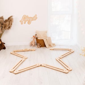 May include: A wooden star-shaped balance beam set on a light wood floor. A woven basket with a blanket and pillow is in the background, along with a wooden sign that says "Abigail". A stuffed giraffe and teddy bear are also in the background.