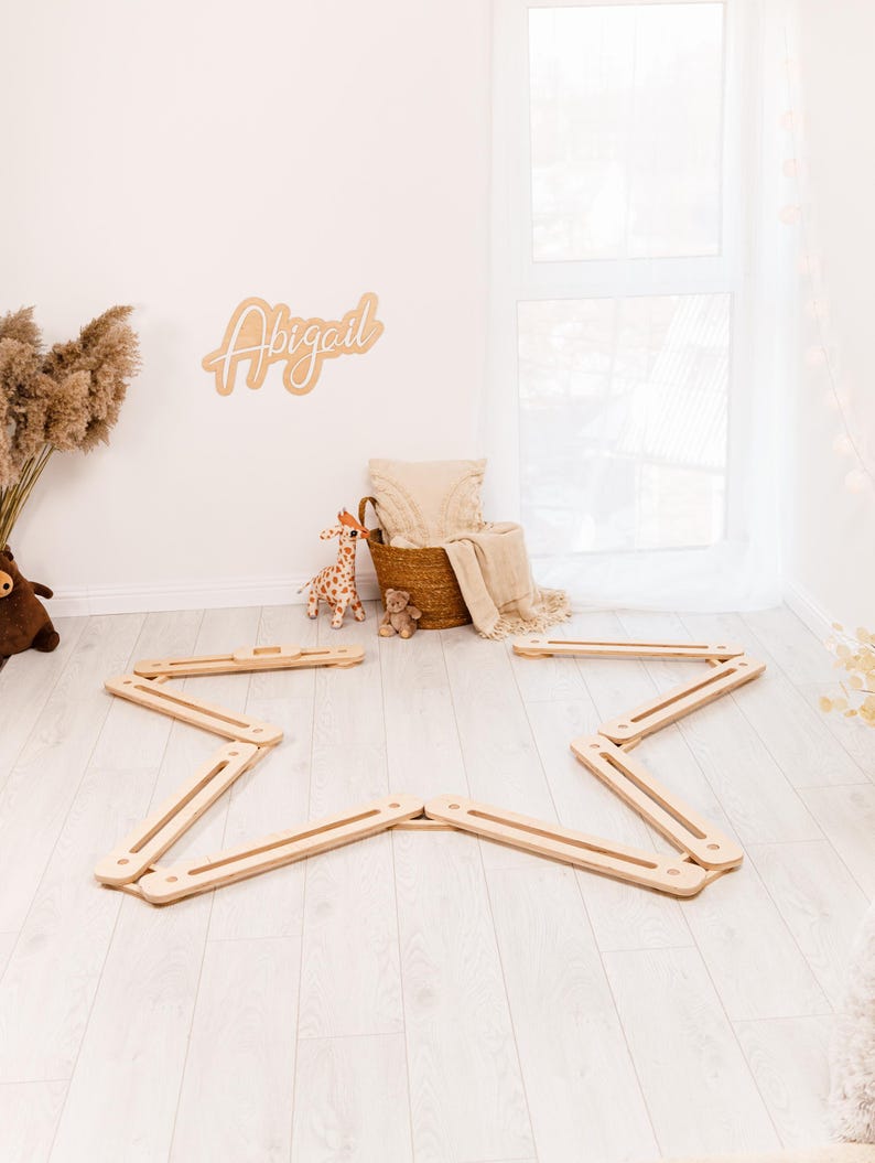May include: A wooden star-shaped balance beam set on a light wood floor. A woven basket with a blanket and pillow is in the background, along with a wooden sign that says "Abigail". A stuffed giraffe and teddy bear are also in the background.