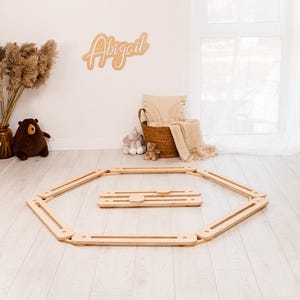 May include: A wooden play gym set on a light wood floor. The set includes an octagonal frame and a smaller rectangular piece. A wooden sign with the name "Abigail" is on the wall. A basket with a blanket and stuffed animals is in the background.