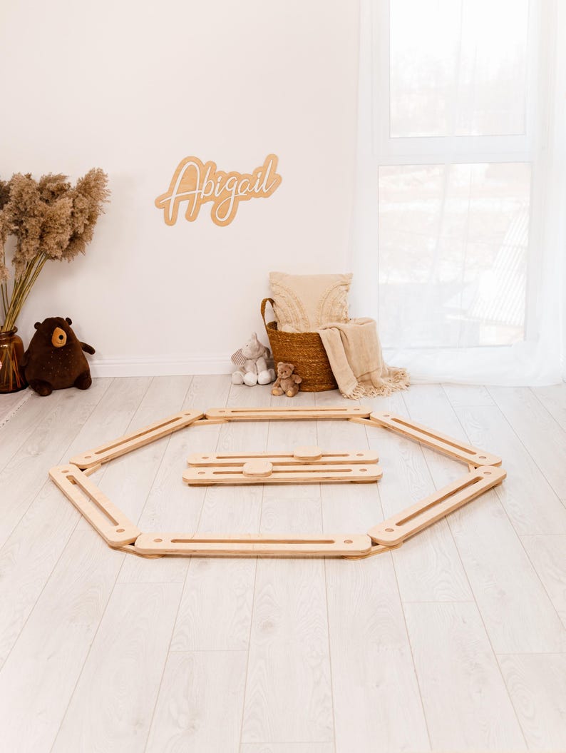 May include: A wooden play gym set on a light wood floor. The set includes an octagonal frame and a smaller rectangular piece. A wooden sign with the name "Abigail" is on the wall. A basket with a blanket and stuffed animals is in the background.