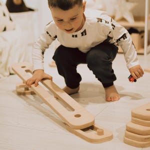 May include: A child playing with a wooden balance board and blocks. The balance board is light brown with two long openings. The child is wearing a white long-sleeve shirt with black geometric shapes and dark trousers.