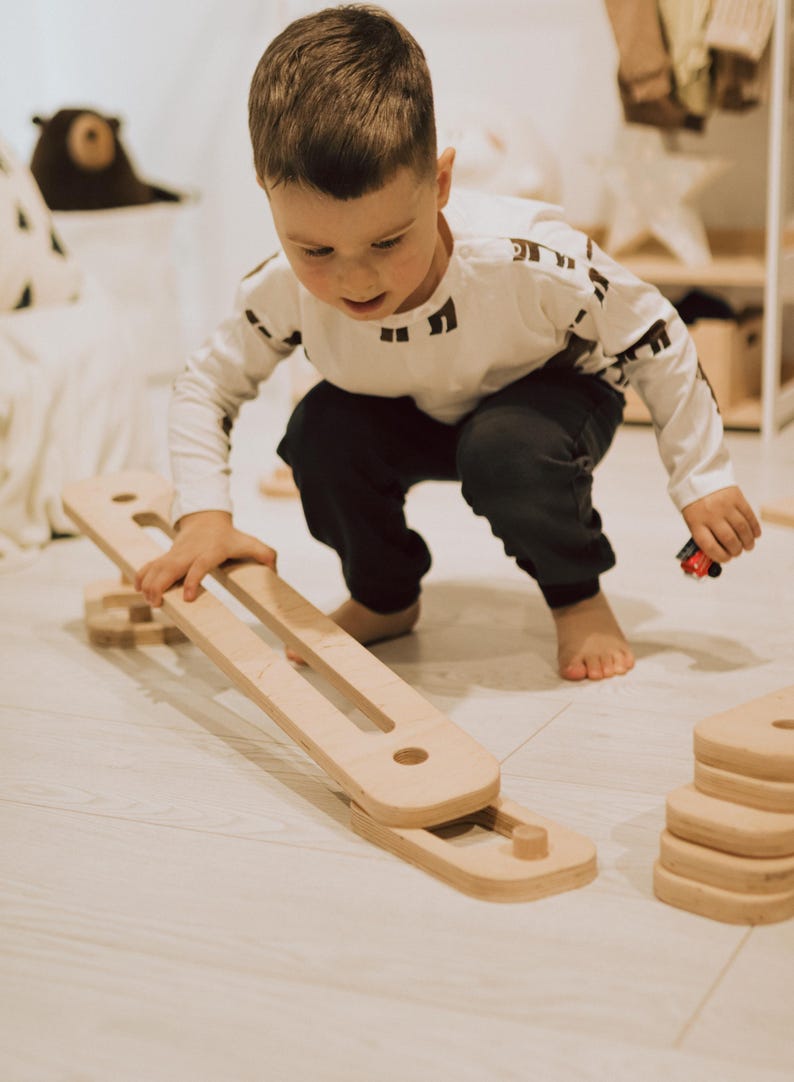 May include: A child playing with a wooden balance board and blocks. The balance board is light brown with two long openings. The child is wearing a white long-sleeve shirt with black geometric shapes and dark trousers.