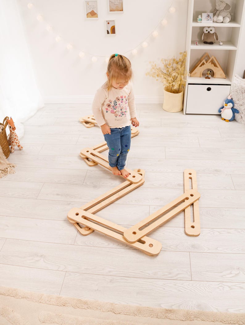 May include: A child stands on a wooden balance beam toy. The toy is made of light-colored wood and has a zig-zag design. The child is wearing a long-sleeved shirt and jeans. The background includes a white wall, a shelf, and decorative items.