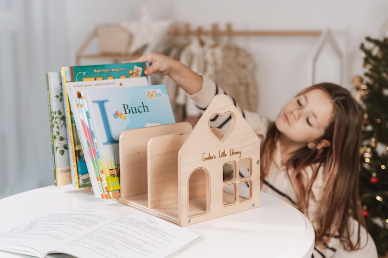 May include: A house-shaped wooden bookshelf with a heart-shaped window and the inscription "Emilia's Little Library." Several books are placed inside the bookshelf. The bookshelf is on a white table. A child is reaching for a book.