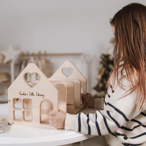 May include: A wooden toy library shaped like a house, featuring a heart-shaped window. The text "Emilia's Little Library" is inscribed on the front. The toy is made of light-coloured wood and has a door and several windows.