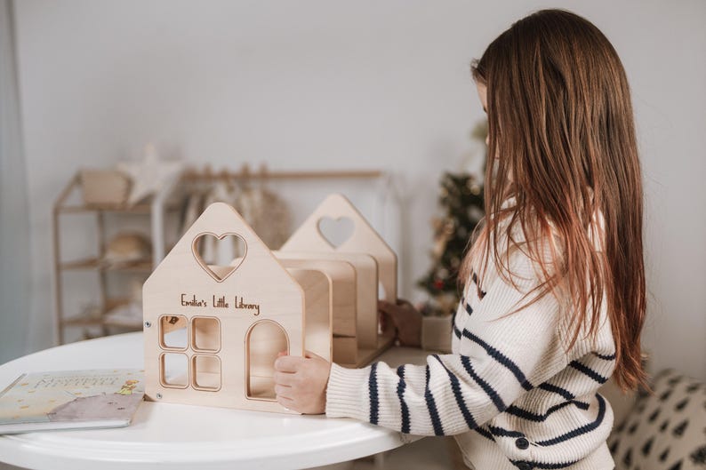 May include: A wooden toy library shaped like a house, featuring a heart-shaped window. The text "Emilia's Little Library" is inscribed on the front. The toy is made of light-coloured wood and has a door and several windows.