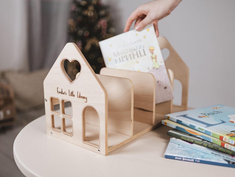 May include: A wooden house-shaped bookshelf with a heart-shaped window and the text "Emilia's Little Library." Several books are visible, with one being placed inside. The design is simple and natural, with a light wood finish. The bookshelf is made of wood.