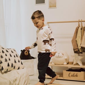May include: A child in a white top with brown bear prints and dark trousers walks on wooden balance beams. A black bear soft toy is in a white basket. A wooden box labelled "Caleb" sits on a shelf. Soft toys and artwork are visible.