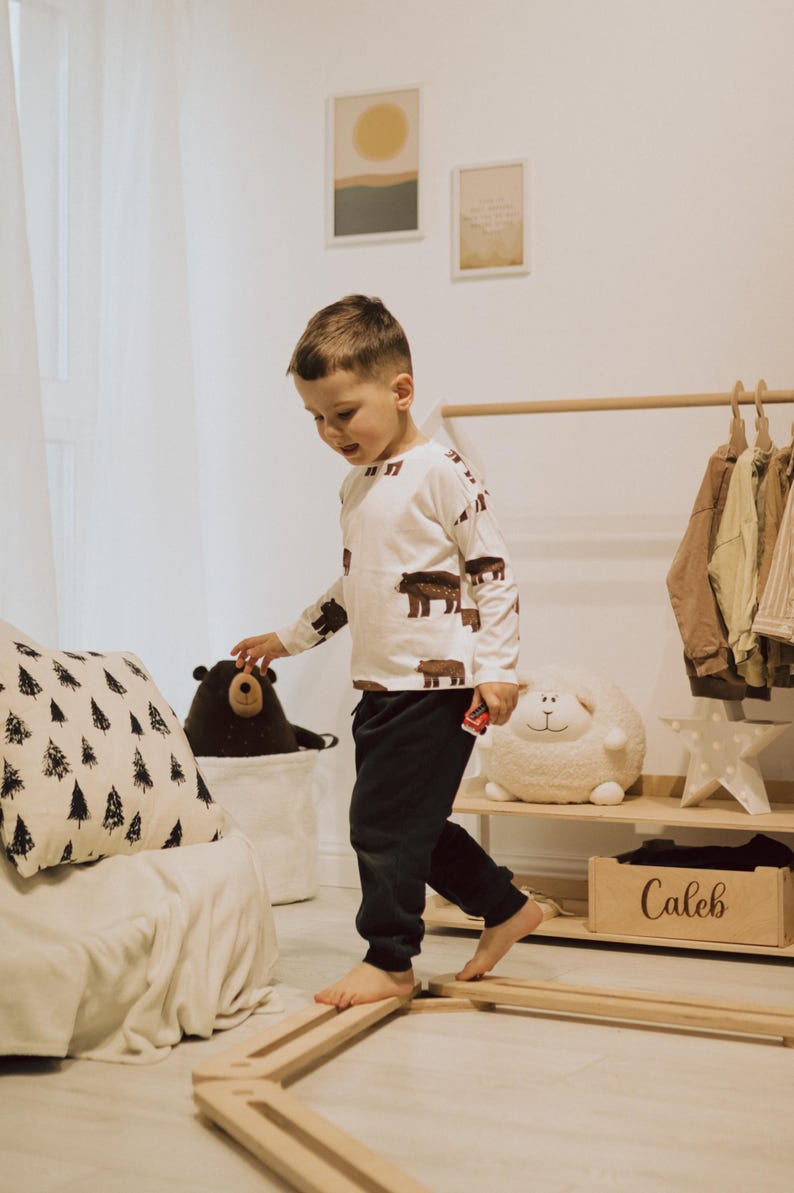 May include: A child in a white top with brown bear prints and dark trousers walks on wooden balance beams. A black bear soft toy is in a white basket. A wooden box labelled "Caleb" sits on a shelf. Soft toys and artwork are visible.