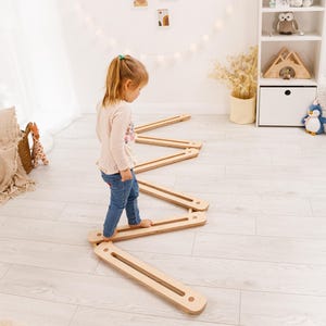 May include: A child is walking on a wooden balance beam set. The set is made of light-coloured wood and arranged in a zig-zag pattern. The room has a neutral colour palette with white walls and light wood flooring. A white shelf with toys is in the background.