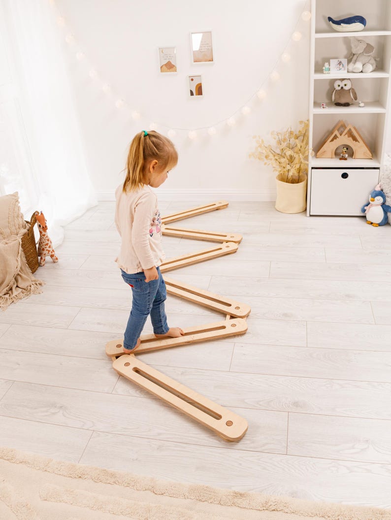 May include: A child is walking on a wooden balance beam set. The set is made of light-coloured wood and arranged in a zig-zag pattern. The room has a neutral colour palette with white walls and light wood flooring. A white shelf with toys is in the background.
