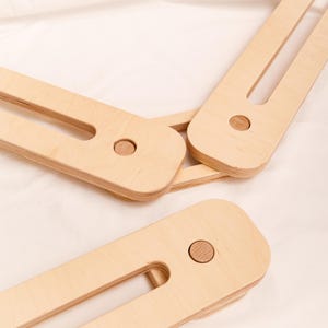 May include: Close-up of several light-coloured wooden pieces, possibly for a frame or support structure. Each piece has a rounded end with a circular wooden insert and a long, rectangular body with a central cutout. The wood grain is visible.