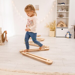 May include: A young child is carefully stepping across a series of wooden balance beams. The beams are light brown and rectangular. The child wears a floral top and blue jeans. The beams are approximately 100 cm long.