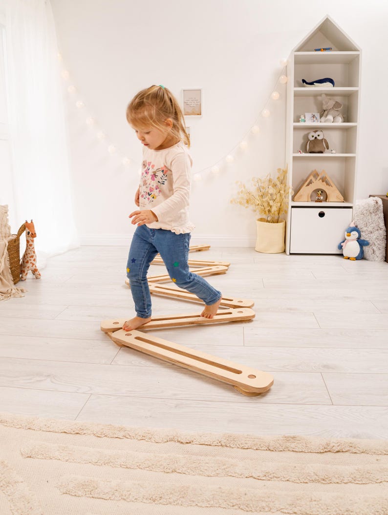May include: A young child is carefully stepping across a series of wooden balance beams. The beams are light brown and rectangular. The child wears a floral top and blue jeans. The beams are approximately 100 cm long.