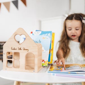 Könnte beinhalten: Ein Bücherregal in Form eines Holzhauses mit herzförmigem Fenster und dem Text "Emilia's Little Library". Das Bücherregal hat mehrere Öffnungen für Bücher. Ein Kind sitzt an einem Tisch mit Büchern.