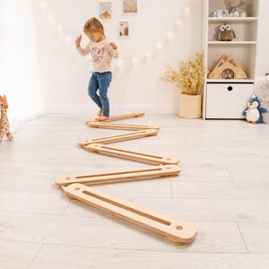 May include: A wooden balance beam toy in a zig-zag pattern on a light wood floor. A young child is walking on the balance beam. The toy is made of light-coloured wood and is designed for children's play and development.