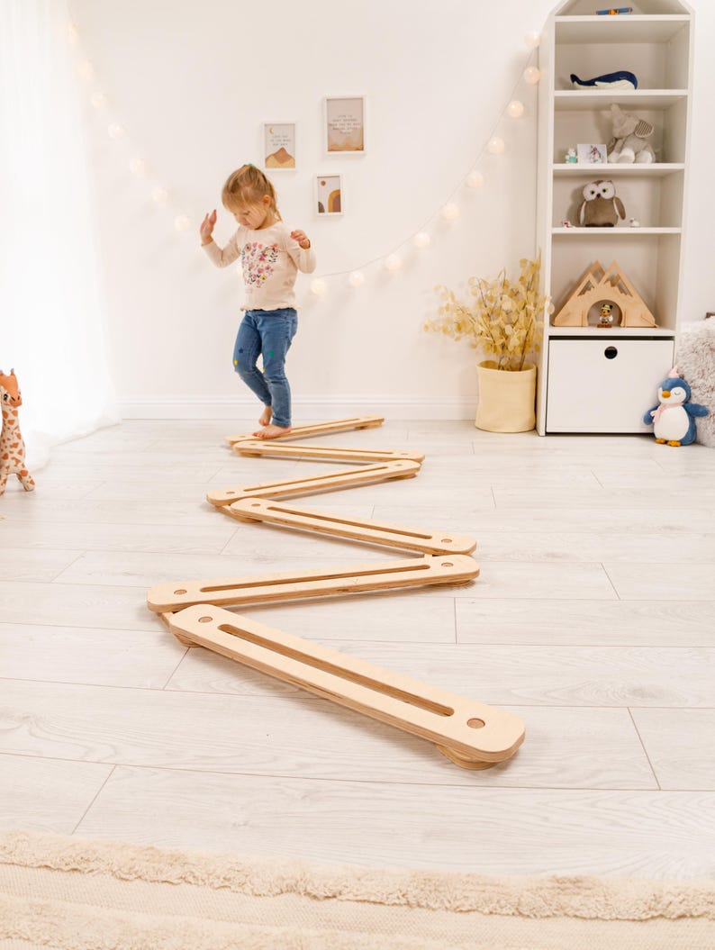 May include: A wooden balance beam toy in a zig-zag pattern on a light wood floor. A young child is walking on the balance beam. The toy is made of light-coloured wood and is designed for children's play and development.