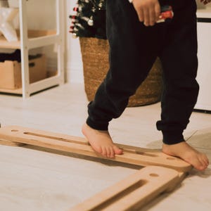 May include: A child walks barefoot on a wooden balance board. The board is made of light-coloured wood and has a rectangular shape with grooves. A small Christmas tree in a woven basket is in the background.