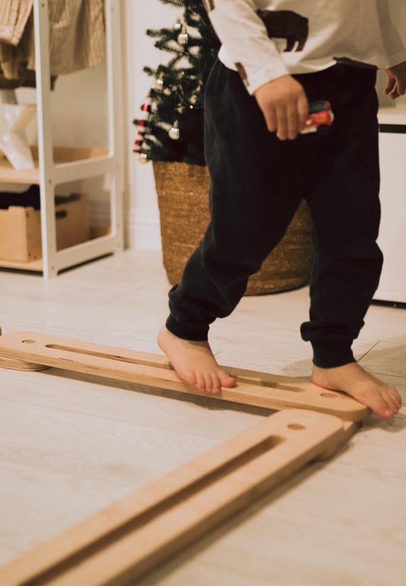 May include: A child walks barefoot on a wooden balance board. The board is made of light-coloured wood and has a rectangular shape with grooves. A small Christmas tree in a woven basket is in the background.