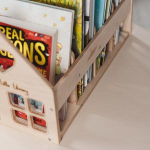 May include: A light wooden house-shaped bookshelf holding a selection of children's books. The bookshelf features a house design with window cutouts and the words "Little Library". The books have bright covers, including titles like "Read with Me".