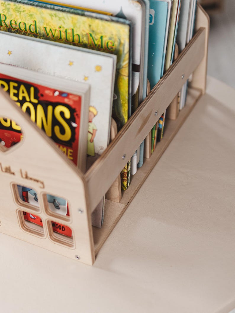 May include: A light wooden house-shaped bookshelf holding a selection of children's books. The bookshelf features a house design with window cutouts and the words "Little Library". The books have bright covers, including titles like "Read with Me".