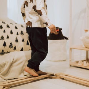 May include: A child's long-sleeved top with brown bear prints, worn with dark blue joggers. The child is walking on a wooden balance beam. A toy car is held in the child's hand. The room has a neutral colour palette.