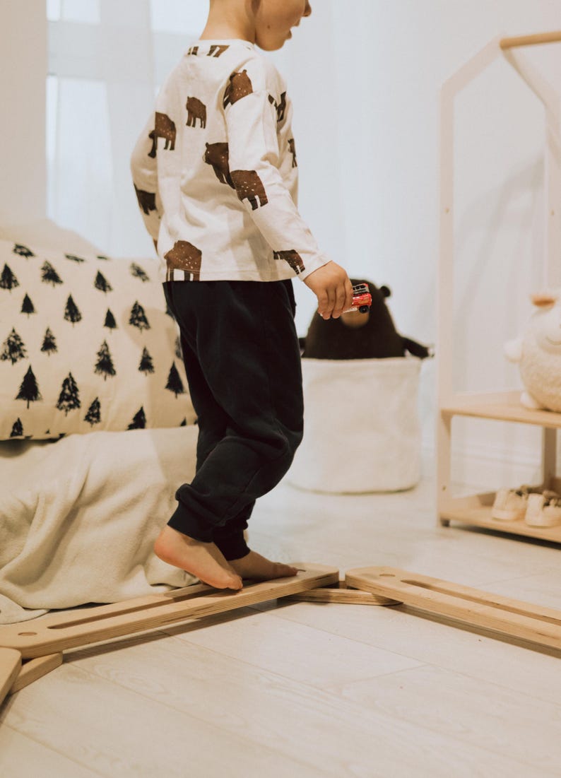 May include: A child's long-sleeved top with brown bear prints, worn with dark blue joggers. The child is walking on a wooden balance beam. A toy car is held in the child's hand. The room has a neutral colour palette.