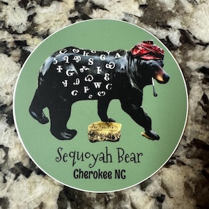 May include: A green circle sticker with a black bear wearing a red bandana and holding a pipe. The bear has white letters on its back that spell out the Cherokee alphabet. The sticker says "Sequoyah Bear Cherokee NC".