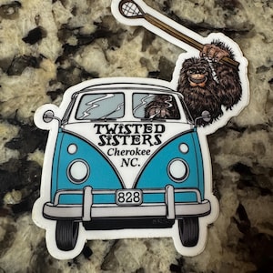 May include: A blue and white sticker of a Volkswagen van with a Bigfoot character driving. The van has the text "Twisted Sisters" and "Cherokee NC" on the side. The van has the number "828" on the front bumper. The Bigfoot character is holding a lacrosse stick.
