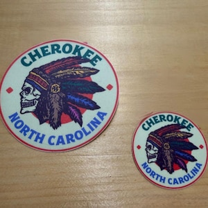 May include: Two round stickers with a Native American skull design. The stickers feature the text "CHEROKEE" and "NORTH CAROLINA" in a circular pattern. The design includes a skull wearing a feathered headdress, with red, blue, and purple accents.