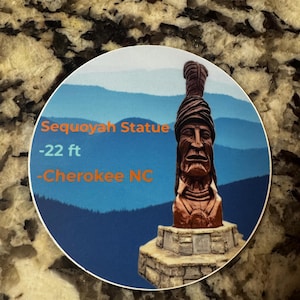 Op de afbeelding: Een bruin houten standbeeld van een indiaanse man met een hoofdtooi, staande op een stenen basis. Het standbeeld is 6,7 meter hoog en bevindt zich in Cherokee, North Carolina. De achtergrond is een blauwe lucht met witte wolken en een bergketen.