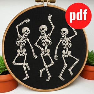 Pode incluir: Uma obra de arte bordada com três esqueletos dançando sobre um fundo preto, em um bastidor de madeira. Os esqueletos são brancos. Um círculo vermelho com a palavra "pdf" está no canto superior direito.