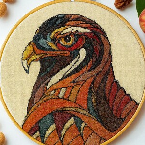 Può includere: Un ricamo a punto croce dettagliato della testa di un falco, con una vibrante tavolozza di colori di marroni, arancioni, rossi e blu. L'opera d'arte è racchiusa in un telaio da ricamo rotondo in legno, che mostra l'occhio e il becco dell'uccello.