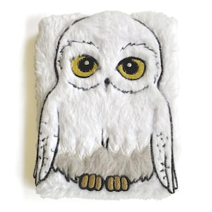May include: A plush, rectangular owl-shaped pillow with a white, fluffy exterior. The owl features large yellow eyes, a gray chest, and black embroidered details. The feet are gold.