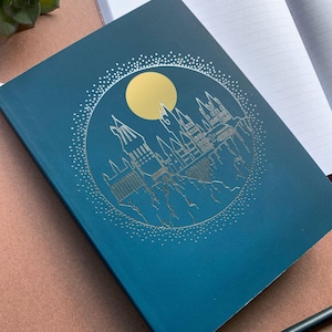 May include: A teal blue notebook with a silver embossed design of Hogwarts castle and a gold moon. The notebook has lined pages.