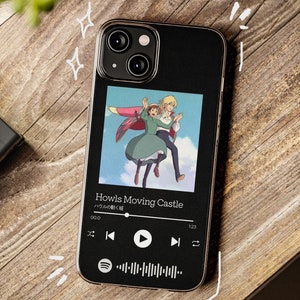 May include: Black phone case featuring a colorful image from "Howl's Moving Castle." The case displays a music player interface with the title of the movie and a Spotify logo. The phone case is on a wooden surface.