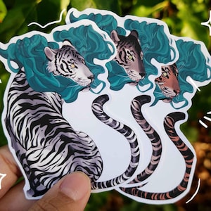 May include: A collection of stickers featuring stylized tiger illustrations. Each sticker showcases a tiger with a teal mane and a unique color palette, including black and white, grey, and brown stripes. The stickers are held against a blurred green background.