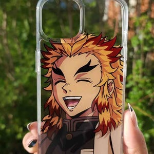 Op de afbeelding: Transparante telefoonhoes met een lachend anime-personage met geel en rood haar. Het personage draagt een zwart uniform met een witte kraag. De hoes wordt tegen een vage groene achtergrond gehouden.