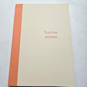 Może przedstawiać: Kremowy notatnik z pomarańczowym grzbietem. Na okładce pomarańczowym kolorem wydrukowano słowa "Trust the process". Notatnik leży płasko na białej powierzchni.