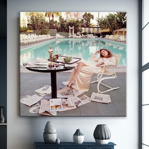 May include: A framed print of a woman lounging by a pool, surrounded by newspapers. A table holds a tea set and an Oscar statue. The scene is set outdoors with palm trees and a pool in the background.