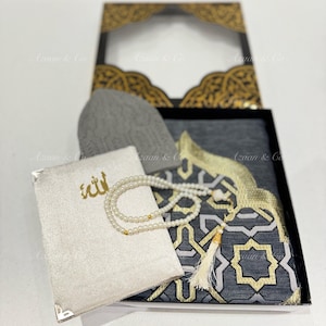 May include: A prayer set in a black box with a gray prayer rug, a white prayer mat with gold Arabic calligraphy, a gray prayer cap, and a white prayer bead necklace.